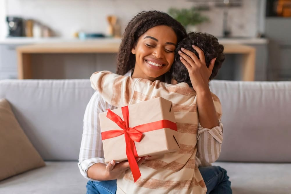 Dia das mães Dia das mães, uma mulher de pele negra, cabelos castanhos, olhos castanhos, jovem, sorrindo ao abraçar seu filho ao granhar um presente de dia das mães.
